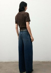 Suéter corto de manga corta marrón y suave, combinado con unos jeans de pierna ancha de color azul oscuro. Cinturón negro con tachuelas plateadas. Fondo minimalista.