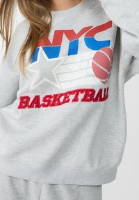 Harmaa huppari, jossa on "NYC" sinisellä ja punaisella, sisältäen koripallografiikan ja "BASKETBALL" paksulla punaisella tekstillä. Pehmeä rakenne.