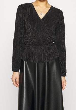 Donna che indossa una camicetta nera plissettata a manica lunga con scollo a V e una gonna in pelle nera, accessoriate con una piccola collana con ciondolo.
