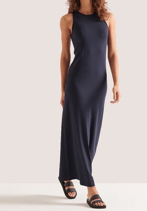 Femme portant une robe longue bleu marine sans manches avec des sandales noires à double brides, debout devant un fond clair uni.