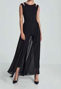 Frau in schwarzem ärmellosem Jumpsuit mit fließendem transparentem Rocküberzug und schwarzen spitzen Schuhen auf einem schlichten hellen Hintergrund.