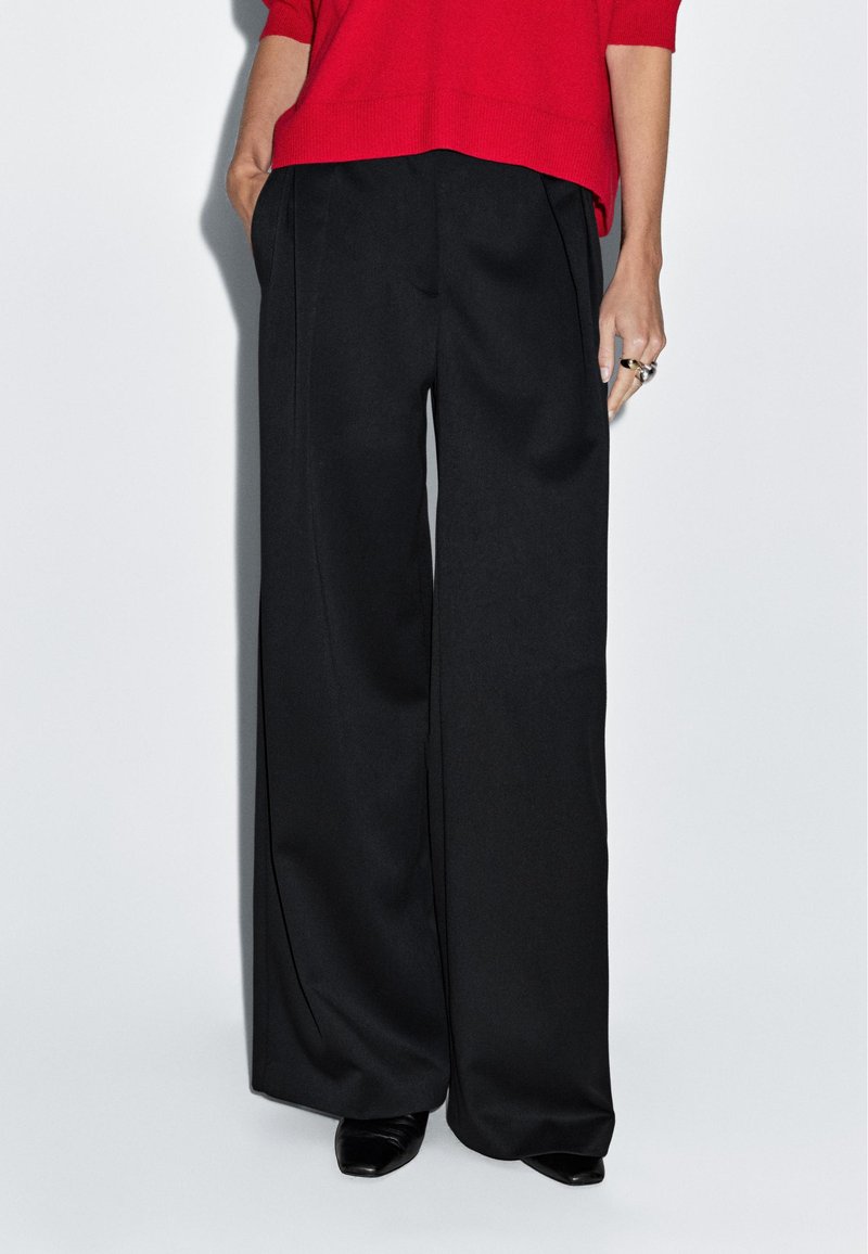 Black wide-leg trousers with a smooth texture and pleated front. Paired with a red knit top and black pointed-toe shoes.