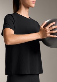 Žena v černém tričku drží černou cvičební kouli v úrovni hrudi na jednolitém pozadí.