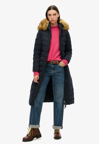 Lange marineblauwe pufferjas met een bont afgewerkte capuchon, gedragen over een roze trui. Kenmerkt brede denimbroeken met manchet en bruine veterschoenen.