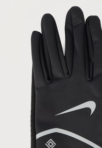 Μαύρο γάντι Nike με ανακλαστικό λευκό λογότυπο Swoosh και γεωμετρική γραμμή σε ανοιχτό φόντο, δάχτυλα τεντωμένα και κοντά το ένα στο άλλο.