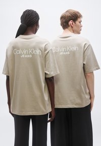 Tricouri bej supradimensionate cu logo-ul "Calvin Klein JEANS" pe spate, realizate din bumbac moale, asortate cu pantaloni negri.