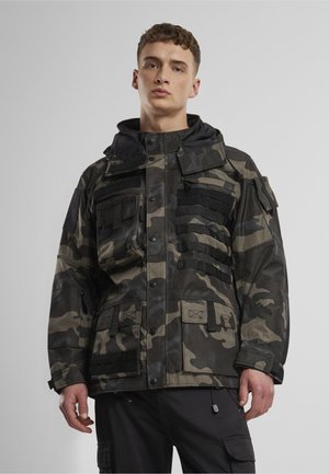 Junger Mann trägt eine schwarze und graue Camouflage-Jacke mit mehreren Taschen und schwarze Cargohosen vor einem einfarbigen Hintergrund.