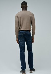 Hombre de pie de espaldas, vistiendo un suéter de manga larga beige, jeans oscuros y zapatillas negras con suelas blancas frente a un fondo gris liso.