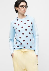 Persoană care poartă ochelari negri și un top cu mâneci lungi, de culoare albastru deschis, cu buline maro, asortat cu pantaloni palid galbeni cu șnur.