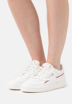 Bílé kožené tenisky s růžovými pruhy, texturovanou gumovou podrážkou a děrovaným detailem na špičce. Součástí jsou tkaničky a nastavitelné polstrování kolem kotníku.