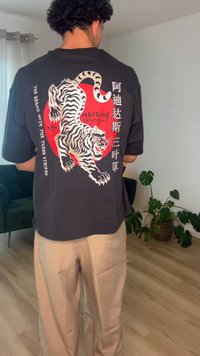 Vyras stovintis patalpose, dėvintis juodą Adidas marškinėlį su baltos tigro grafika ir raudonu apskritimu, mūvi smėlio spalvos kelnes, fone matyti augalai ir baldai.