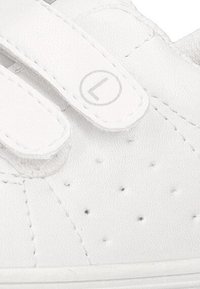 Zapatilla blanca con detalles perforados, correas de velcro ajustables, material de imitación de cuero y un logo circular en el lateral.