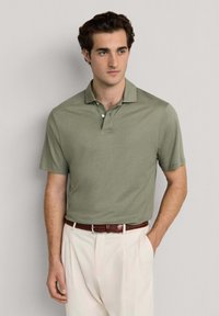 Młody mężczyzna o ciemnych włosach, ubrany w oliwkowozieloną koszulkę polo z krótkim rękawem, włożoną do kremowych spodni z brązowym plecionym paskiem.