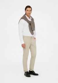 Helle graue Hose, weißes Hemd und ein brauner Strickpullover über die Schultern gelegt. Schwarze Dressschuhe runden das Outfit ab.