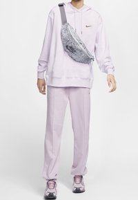 Ljuslila hoodie och sweatpants, tillsammans med en grå fanny pack med ormskinnsmönster. Rosa sneakers med grå detaljer fulländar outfiten.