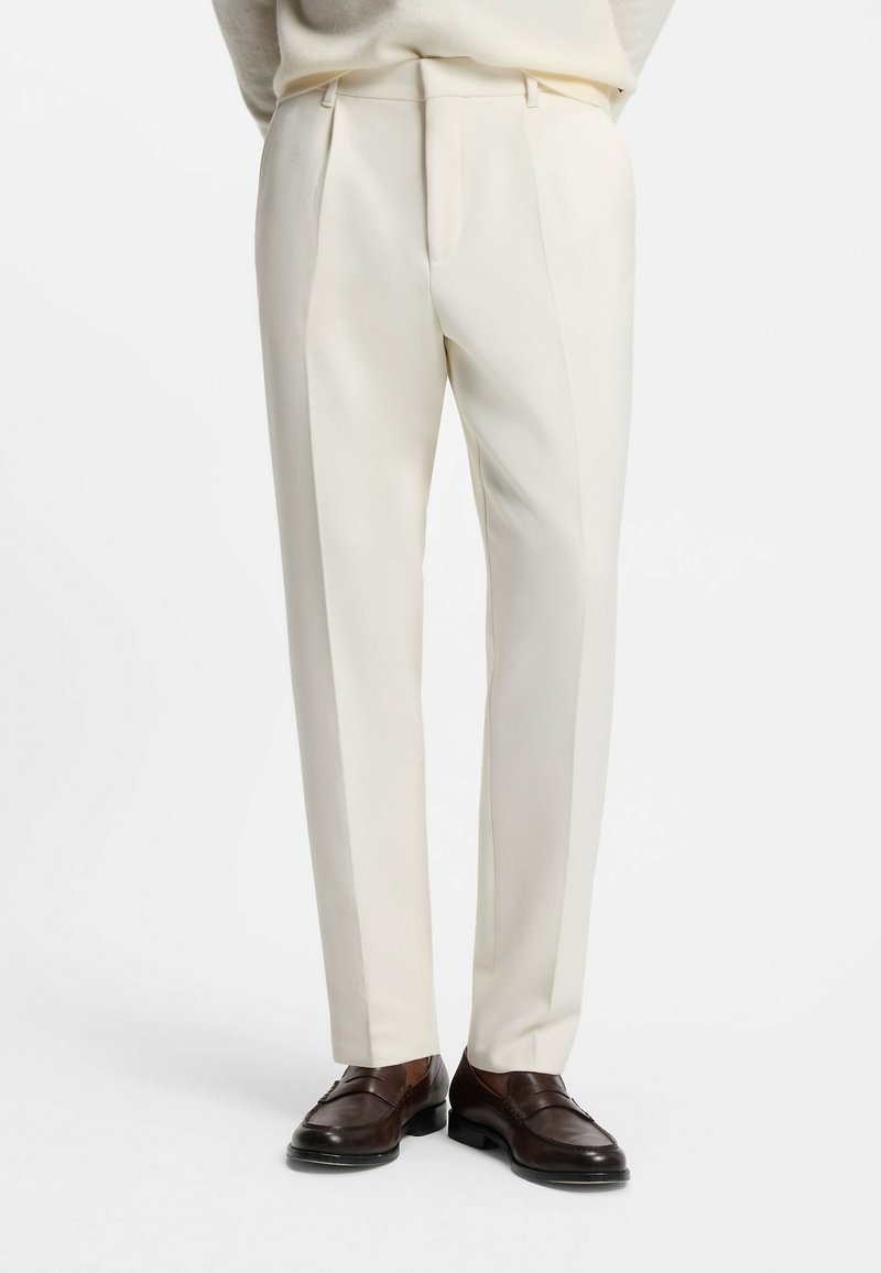 Homme portant un pantalon sur mesure de couleur crème et des mocassins en cuir marron foncé, debout sur un fond blanc.