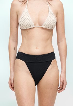 Γυναίκα που φοράει κρεμ πλεκτό μπικίνι τοπ και μαύρο μαγιό με ψηλή μέση και ανάγλυφα λουλουδένια σχέδια, όρθια μπροστά σε ανοιχτό φόντο.