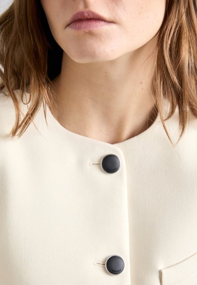 Blazer de couleur crème au toucher lisse, avec un col rond, deux boutons noirs et un détail de poche unique.