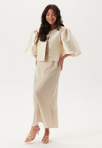 Beige cropped jakke med puffærmer, knapdetaljer og en matchende lang kjole. Glat stof med en subtil glans. Strappy klare hæle.