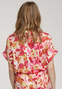 Chemise à manches courtes en tissu léger et texturé avec un motif floral dans des nuances de rose, orange et crème. Comprend une poche au dos.