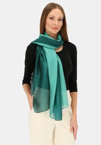 Szalik w kolorze zielonkawoniebieskim wykonany z przezroczystej tkaniny, z gradientowym wzorem. Noszony luźno na szyi, na czarnej bluzce, z swobodnie zwisającymi końcami.