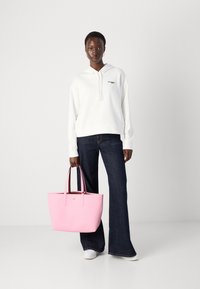 Vit hoodie med grön logotyp, mörkblå utsvängda jeans, rosa tygväska med mörkare handtag, vita sneakers. Enkel, avslappnad outfit.