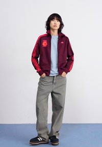 Bordeaux sportovní bunda s červenými pruhy, logem a výšivkou. Svetle modré tričko pod ní. Šedé volné kalhoty a černé boty s bílými pruhy.