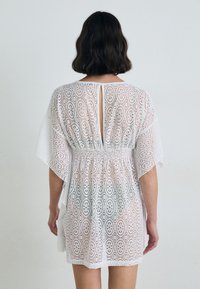 White lace dress features an open back, short sleeves, and a gathered waistline. Decorative oval cutouts create a patterned texture.