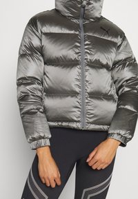 Grå puffjacka med en blänkande yta, dragkedjeslutning, huva och elastiska ärmmuddar. Bärs över svarta leggings med ljusgrå detaljer.