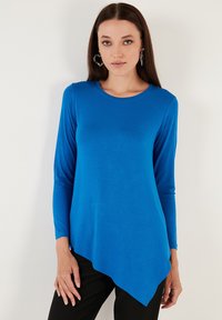 Maglietta a maniche lunghe di un blu brillante, con scollo rotondo e orlo asimmetrico. Il materiale è liscio e leggermente aderente.