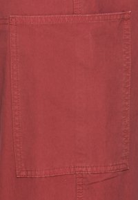 Tela roja con una superficie texturizada, que presenta un gran bolsillo rectangular en la parte frontal. Costuras visibles a lo largo de los bordes.