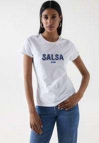 Biała bawełniana koszulka z niebieskim haftowanym napisem "SALSA JEANS", w połączeniu z niebieskimi dżinsami. Krótkie rękawy, okrągły dekolt.