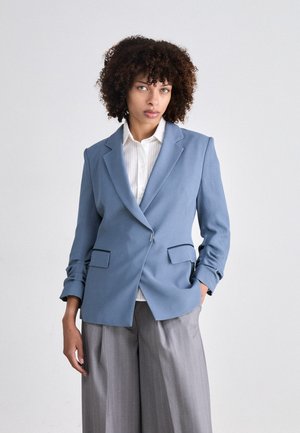 Světle modrý blazer s jedním knoflíkem, dvěma předními kapsami a ohrnutými manžetami, doplněný bílou košilí a šedými pruhovanými kalhotami.
