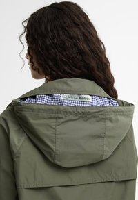 Γυναίκα με σγουρά μαλλιά που φοράει μια ελιάς πράσινη τζάκετ με κουκούλα, δείχνοντας τη μπλε καρό εσωτερική επένδυση και την ετικέτα ρούχων μέσα στον γιακά.