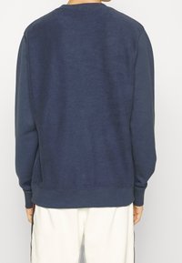 Person klädd i en marinblå fleece-tröja och vita byxor, vänd bort från kameran mot en enfärgad bakgrund.