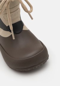 Botte en caoutchouc brun avec une semelle texturée, des accents beige et noir, et un détail de lacet tressé en haut.