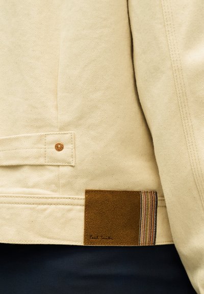 Μπεζ τζιν μπουφάν με κουμπωτή πίσω τσέπη και καφέ σουέντ πατς με διακριτικές πολύχρωμες ρίγες και μικρό λογότυπο.