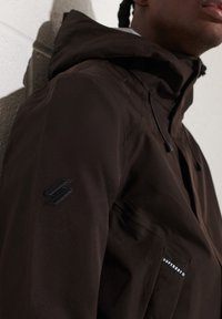 Brązowa wodoodporna kurtka z kapturem, z czarną naszywką z logo na rękawie i kieszenią na suwak. Gładka faktura i stylowy design.