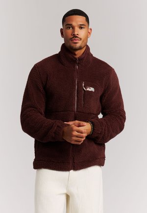 Hombre de pie con las manos entrelazadas, vistiendo una chaqueta de forro polar marrón con un bolsillo con cremallera y pantalones blancos sobre un fondo neutro.