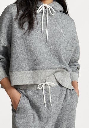 Personne portant un sweat à capuche gris clair et un pantalon de survêtement assorti avec des cordons blancs, mains dans les poches, sur un fond uni.