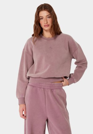 Młoda kobieta o długich brązowych włosach, ubrana w mauve'owy (fioletoworóżowy) sweter i dopasowane spodnie dresowe, stojąca z jedną ręką w kieszeni.