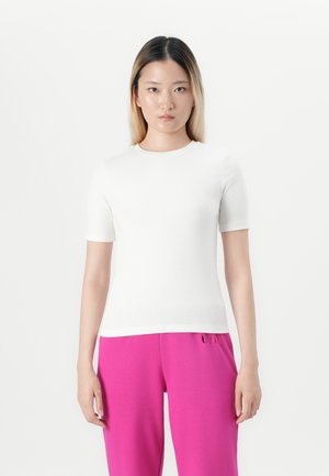 Donna con capelli biondi lisci e lunghi fino alle spalle, che indossa una maglietta bianca semplice a maniche corte e pantaloni rosa brillante, in piedi davanti a uno sfondo bianco.