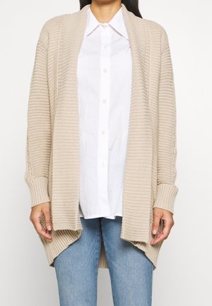 Femme portant un cardigan beige côtelé ouvert sur le devant, sur une chemise blanche boutonnée et un jean bleu clair, debout devant un fond blanc.