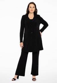 Schwarzes Blazer-Kleid mit langen Ärmeln und Knopfleiste, kombiniert mit schwarzen, weit geschnittenen Hosen und hohen Schuhen. Glatte Stoffoberfläche.