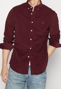 Vínová košile na knoflíky z manšestru s rozšířeným límcem a drobným logem v kombinaci se světle modrými roztrhanými džínami.