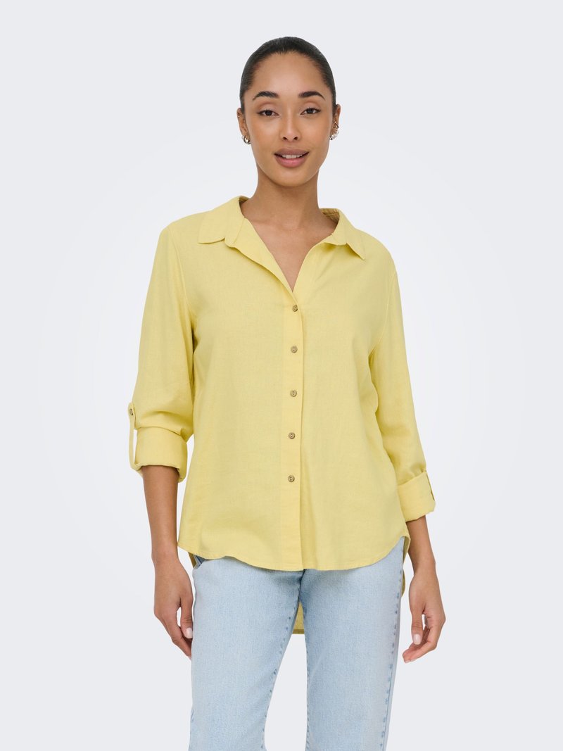 Jeune femme portant une chemise jaune boutonnée aux manches retroussées et un jean bleu clair, debout devant un fond clair uni.