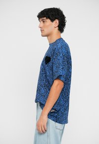 Zila leopardu raksta T-kreklam ir īsas piedurknes, apaļa kakla izgriezums un melna sirsniņas aplikācija uz krūšu daļas. Mīksta auduma, brīvā piegriezumā.