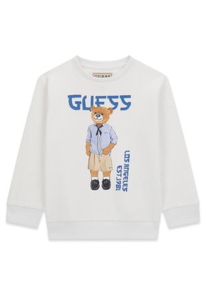 Biała bluza z nadrukiem misia w niebieskiej koszuli, beżowych szortach, czarnych butach oraz niebieskim napisem "GUESS" i "LOS ANGELES EST. 1981".