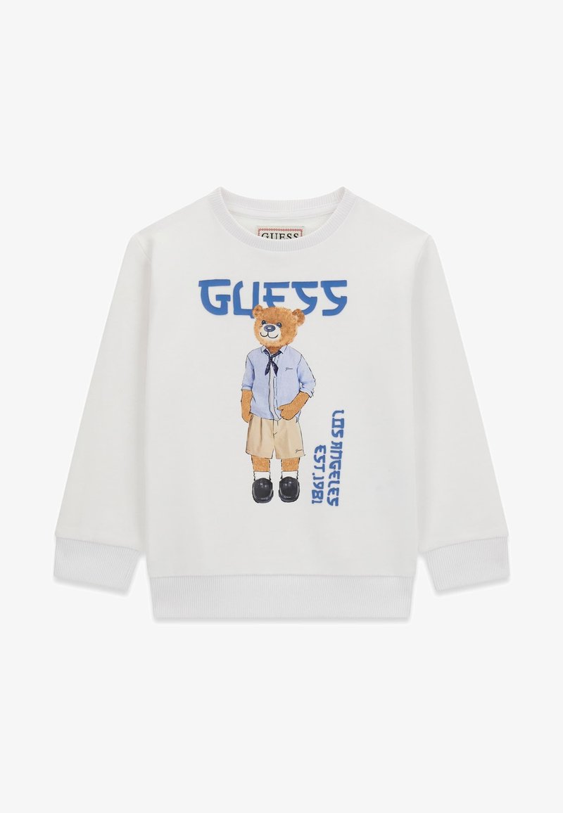 Sweat-shirt blanc avec un ours en peluche portant une chemise bleue, un short beige, des chaussures noires, avec le texte bleu "GUESS" et "LOS ANGELES EST. 1981".
