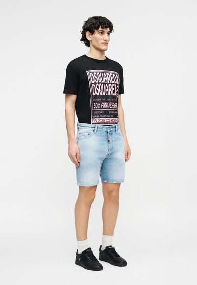 Jeune homme debout portant un t-shirt graphique noir, un short en jean bleu clair, des chaussettes blanches et des baskets noires sur un fond uni.